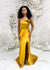 Bridesmaid wearing a modern strapless fitted bridesmaid dress with stretch. The dress has a slit at the leg, and curved architectural detailing at the neck, and is shown in a golden vibrant yellow on a black model.