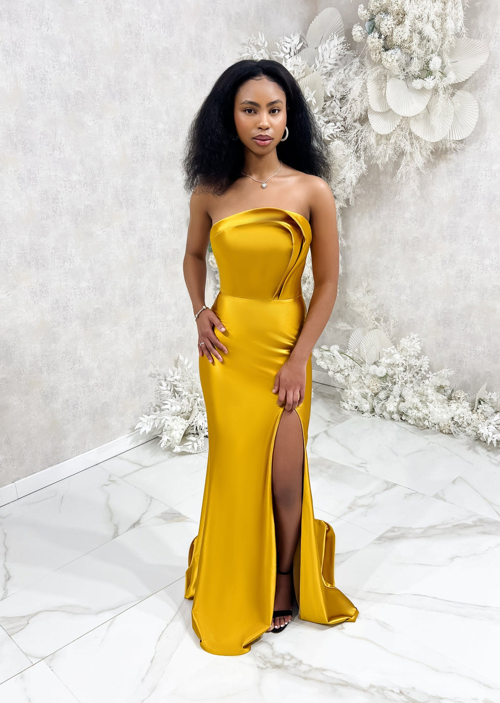Bridesmaid wearing a modern strapless fitted bridesmaid dress with stretch. The dress has a slit at the leg, and curved architectural detailing at the neck, and is shown in a golden vibrant yellow on a black model.