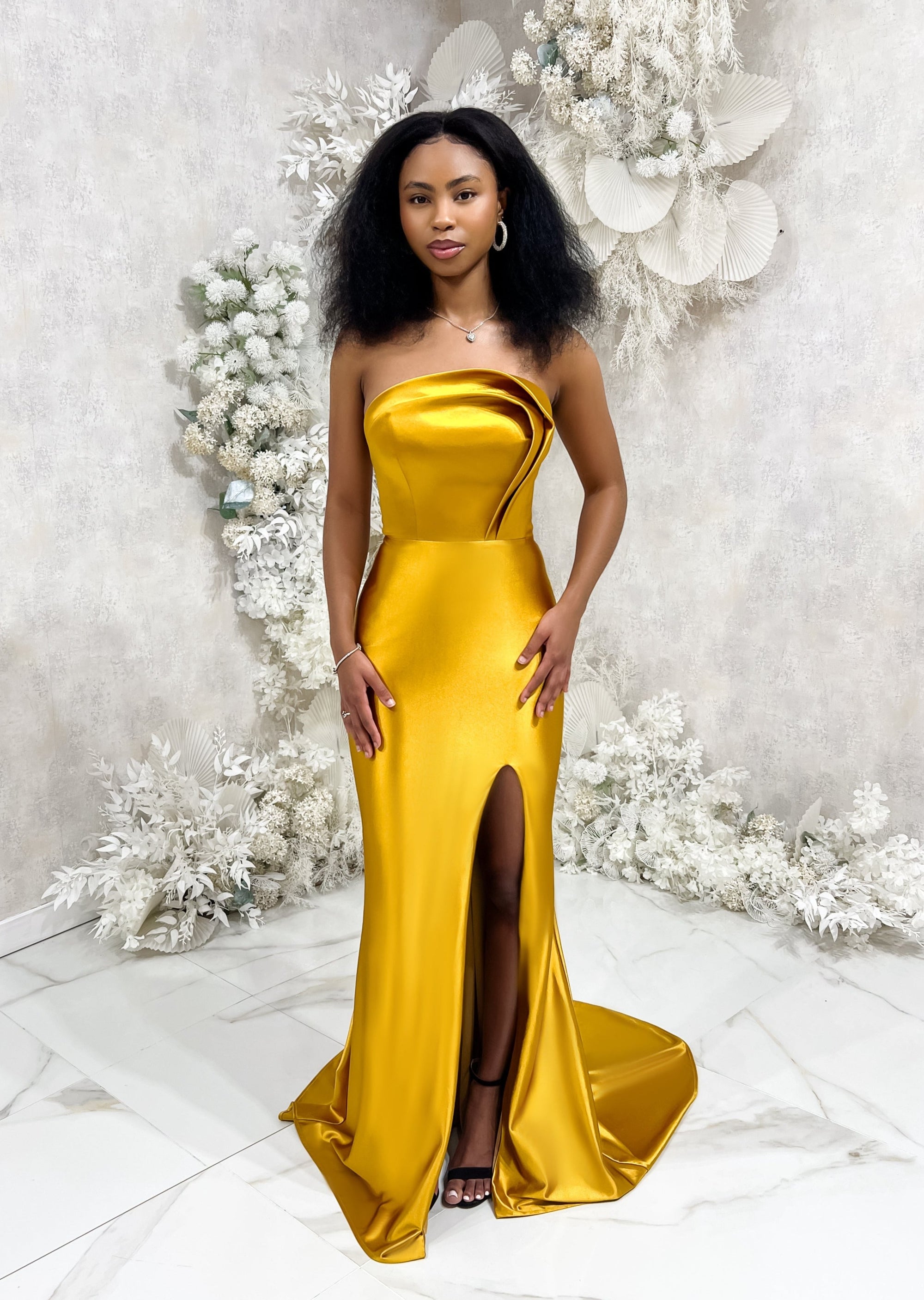 Bridesmaid wearing a modern strapless fitted bridesmaid dress with stretch. The dress has a slit at the leg, and curved architectural detailing at the neck, and is shown in a golden vibrant yellow on a black model.