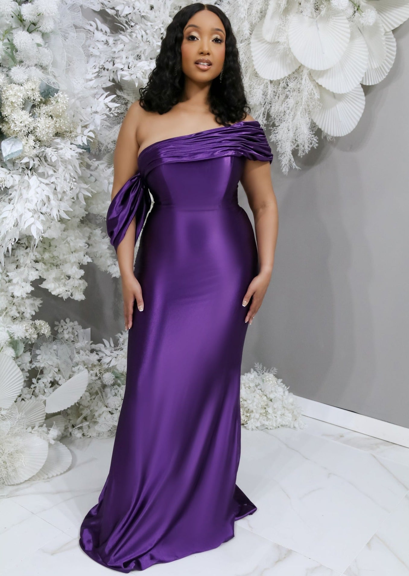 Plus size bridesmaid in asymmetrical fitted stretch satin bridesmaid dress with draping across the neckline continuing to an off-the-shoulder strap. This style is designed to flatter and complement all body types. Shown in jewel tone on a black curvy model.