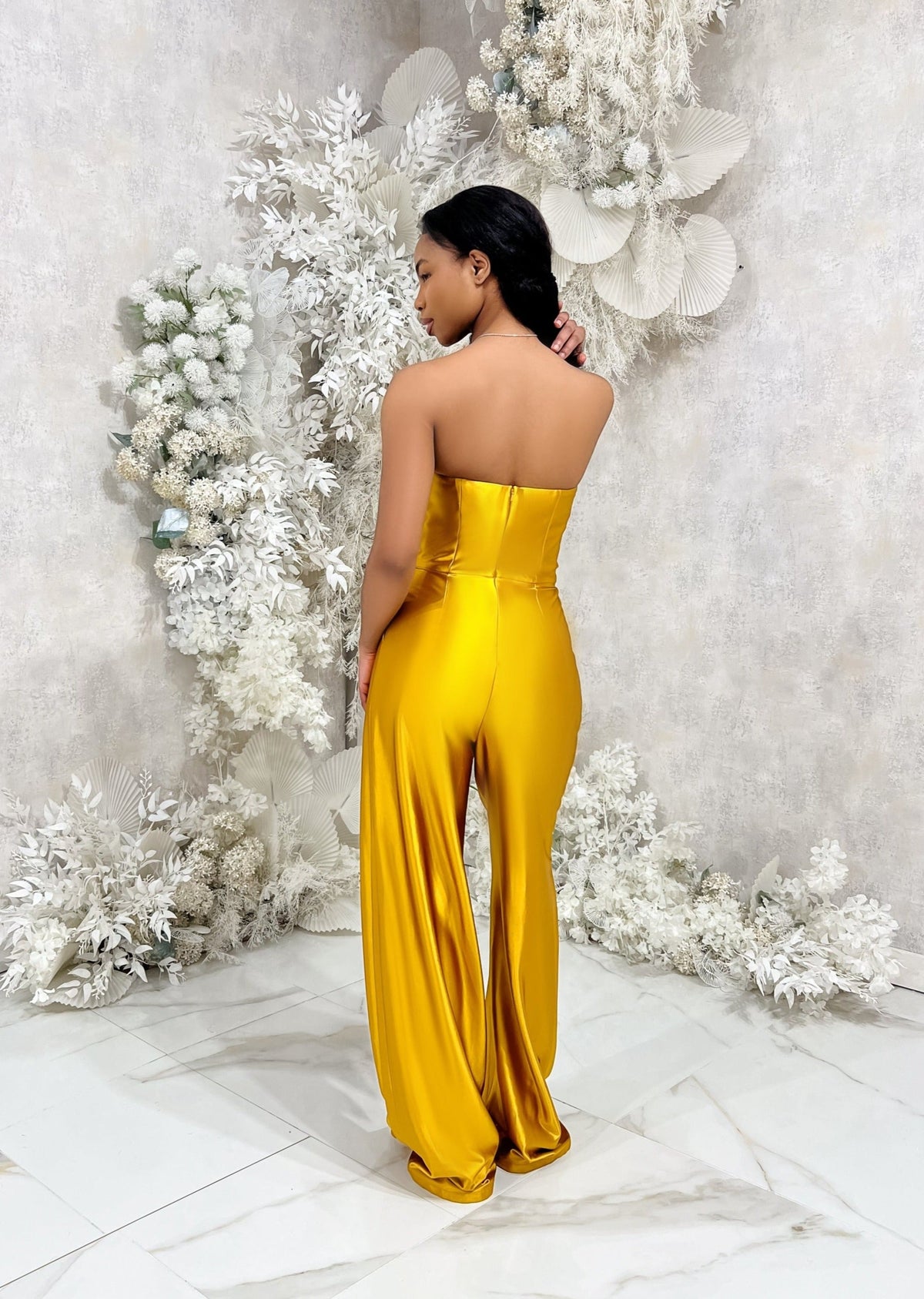 Bridesmaid wearing a modern strapless fitted bridesmaid jumpsuit with stretch. Style has a curved architectural detailing at the neck and is shown in a golden vibrant yellow on a black model.