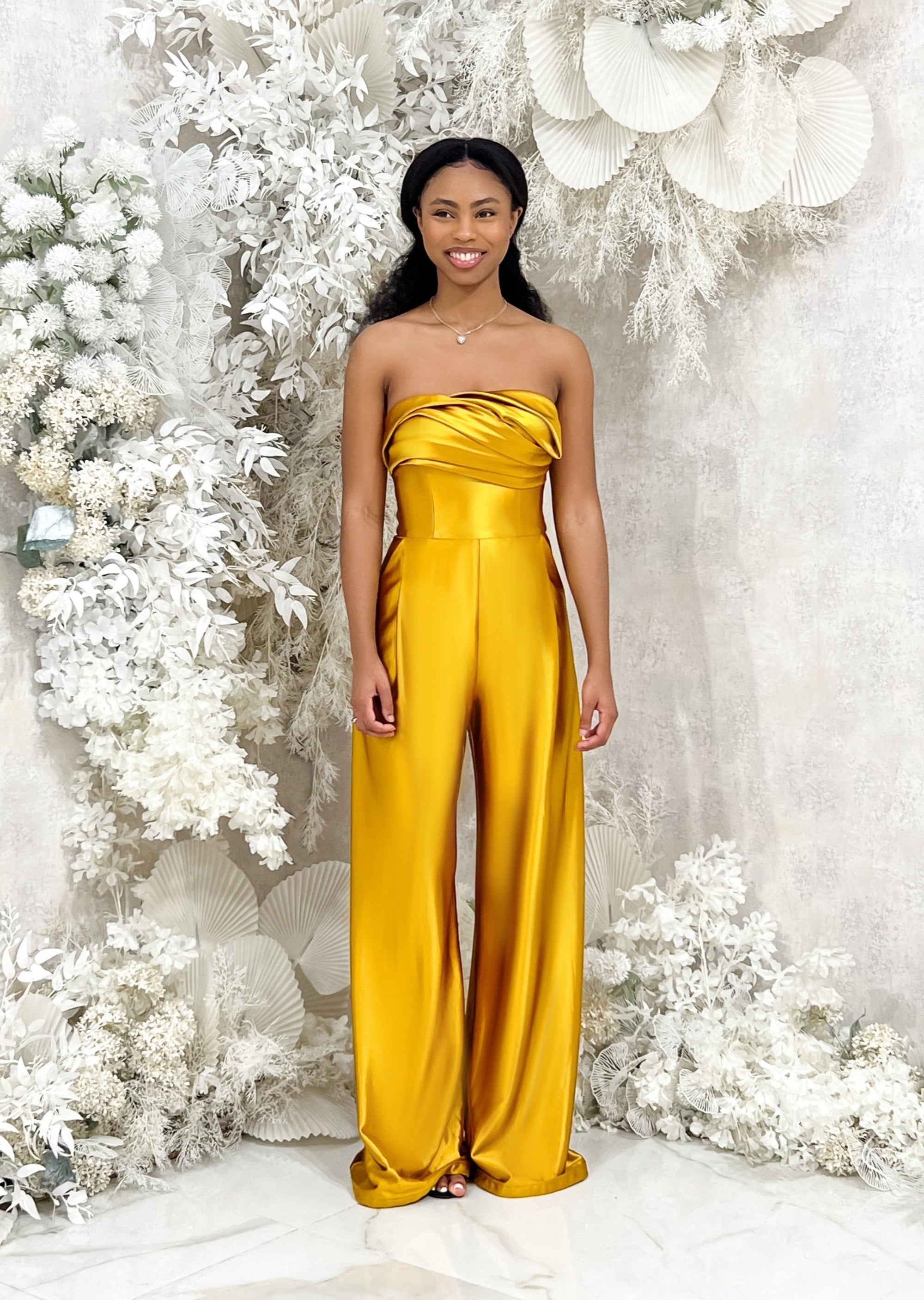 Bridesmaid wearing a modern strapless fitted bridesmaid jumpsuit with stretch. Style has a curved architectural detailing at the neck and is shown in a golden vibrant yellow on a black model.