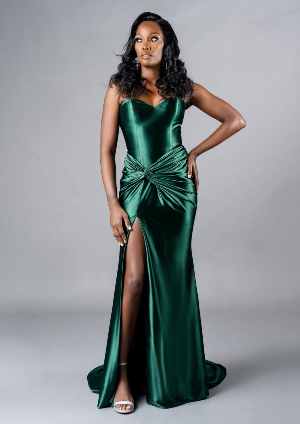 Curvy Bridesmaid wearing a strapless fitted bridesmaids dress with stretch. Dress has a slit at the leg, twisted detailing at the hip, and is shown in emerald green on a black model.