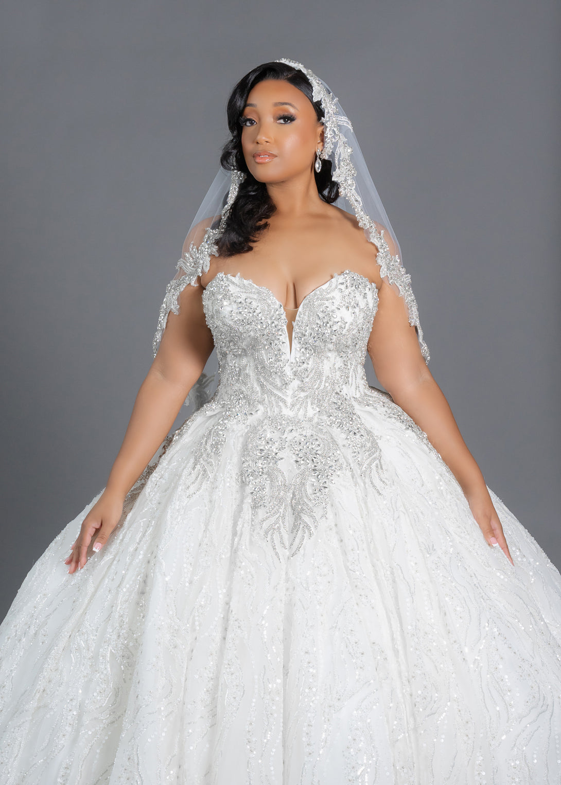 Beautiful finger-tip bridal veil with bling border designed for brides seeking a dramatic addition to their wedding look. Featuring sparkling crystal appliques this veil provides bling along the entire edge. Available in white, diamond white, and ivory with optional blusher. Shown on plus size bride wearing beaded ballgown.