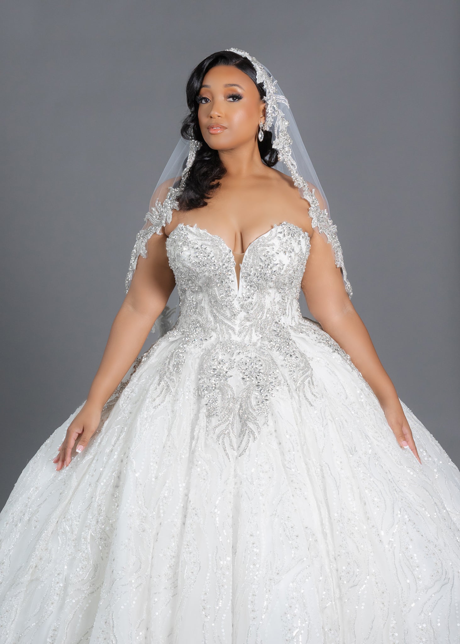 Beautiful finger-tip bridal veil with bling border designed for brides seeking a dramatic addition to their wedding look. Featuring sparkling crystal appliques this veil provides bling along the entire edge. Available in white, diamond white, and ivory with optional blusher. Shown on plus size bride wearing beaded ballgown.