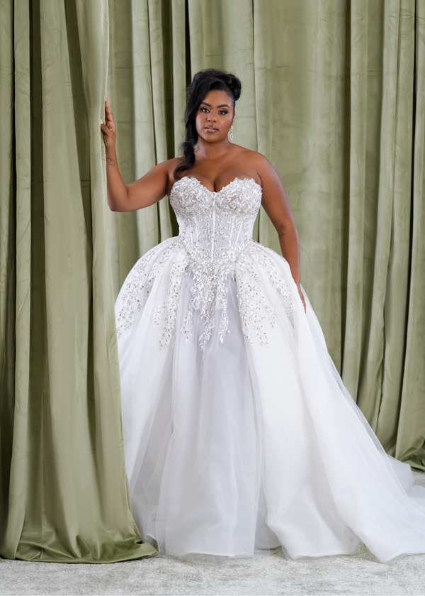 Plus size Black bride wearing a strapless ballgown wedding dress with a sheer corset bodice, exposed boning, crystal appliqué, and full organza skirt, designed by Black bridal brand Pantora.