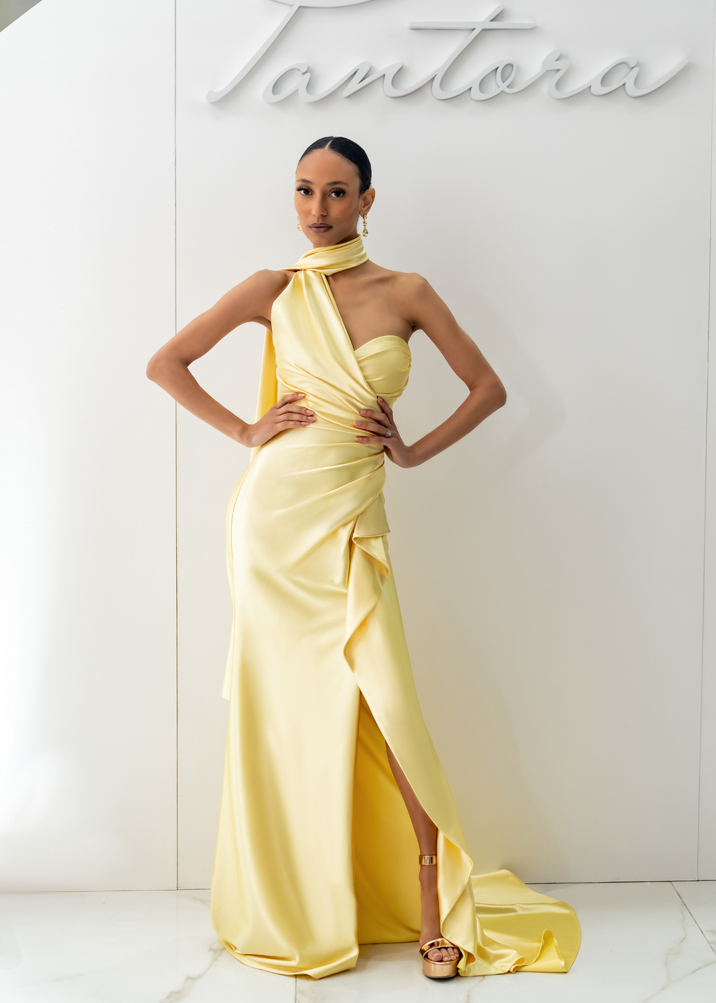 A woman wearing a yellow evening gown with a dramatic scarf detail around the neck and asymmetrical draping. The gown features a sculpted waistline and floor-length design.
