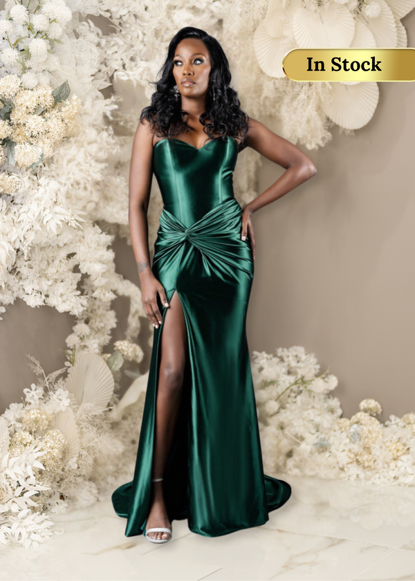 Curvy Bridesmaid wearing a strapless fitted bridesmaids dress with stretch. Dress has a slit at the leg, twisted detailing at the hip, and is shown in emerald green on a black model.