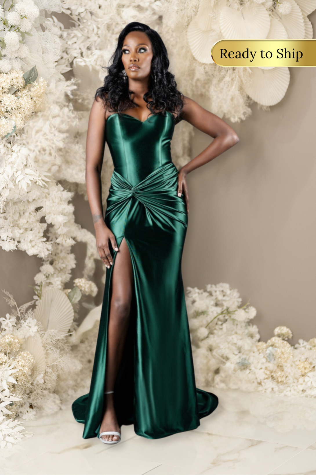 Curvy Bridesmaid wearing a strapless fitted bridesmaids dress with stretch. Dress has a slit at the leg, twisted detailing at the hip, and is shown in emerald green on a black model.