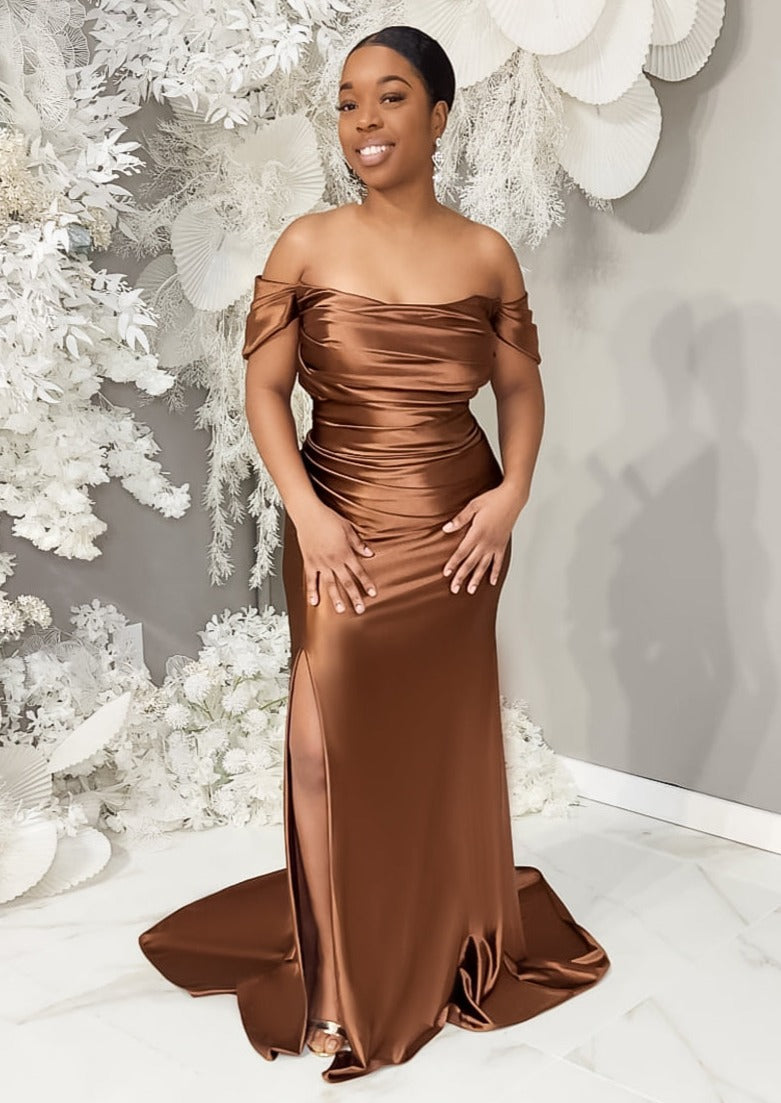 Petite black model wearing a fitted stretch satin bridesmaid dress with off-the-shoulder detailing, ruching, and slit on curvy model