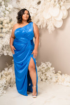 Plus size bridesmaid in asymmetrical column stretch satin bridesmaid dress with pleats and ruching throughout. This style has a slit at the leg and is designed for flatter and comfort all body types. Shown in a bright blue on a black model.