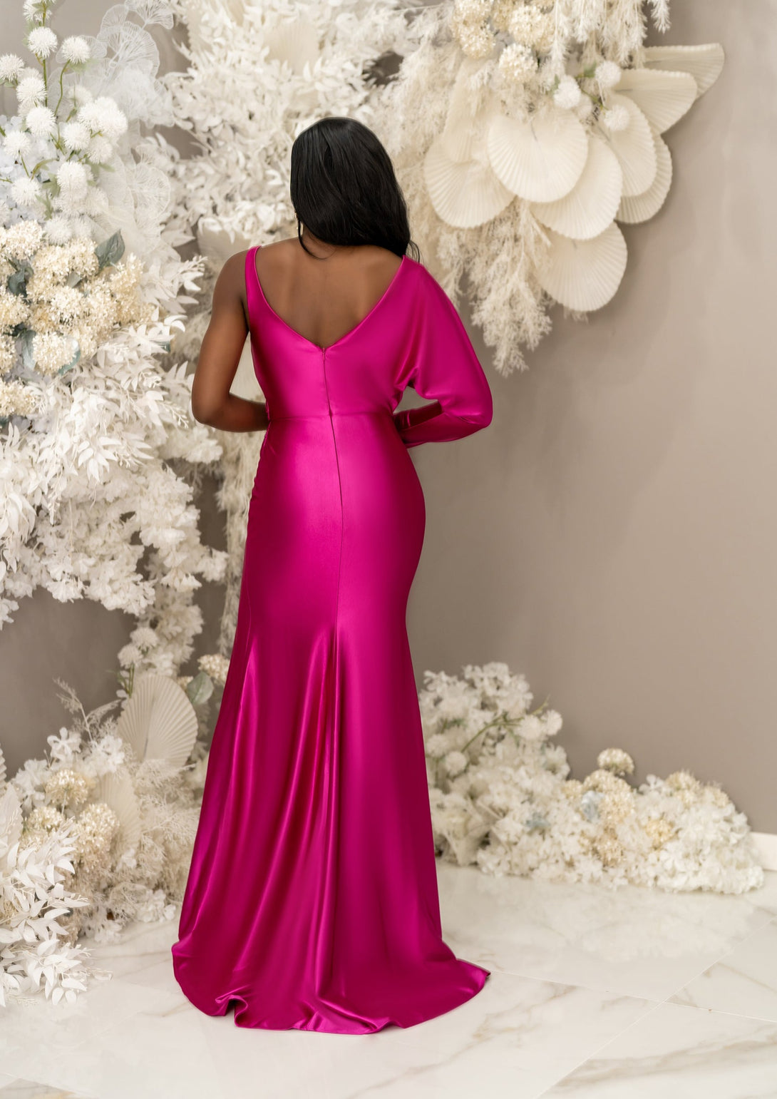 Jewel tone pink fitted V-Neck bridesmaid dress with an asymmetrical Dolman sleeve, has gathering and gentle draping. Shown on a petite black model, this style features a leg slit and is made in stretch satin to flaunt curves - back view