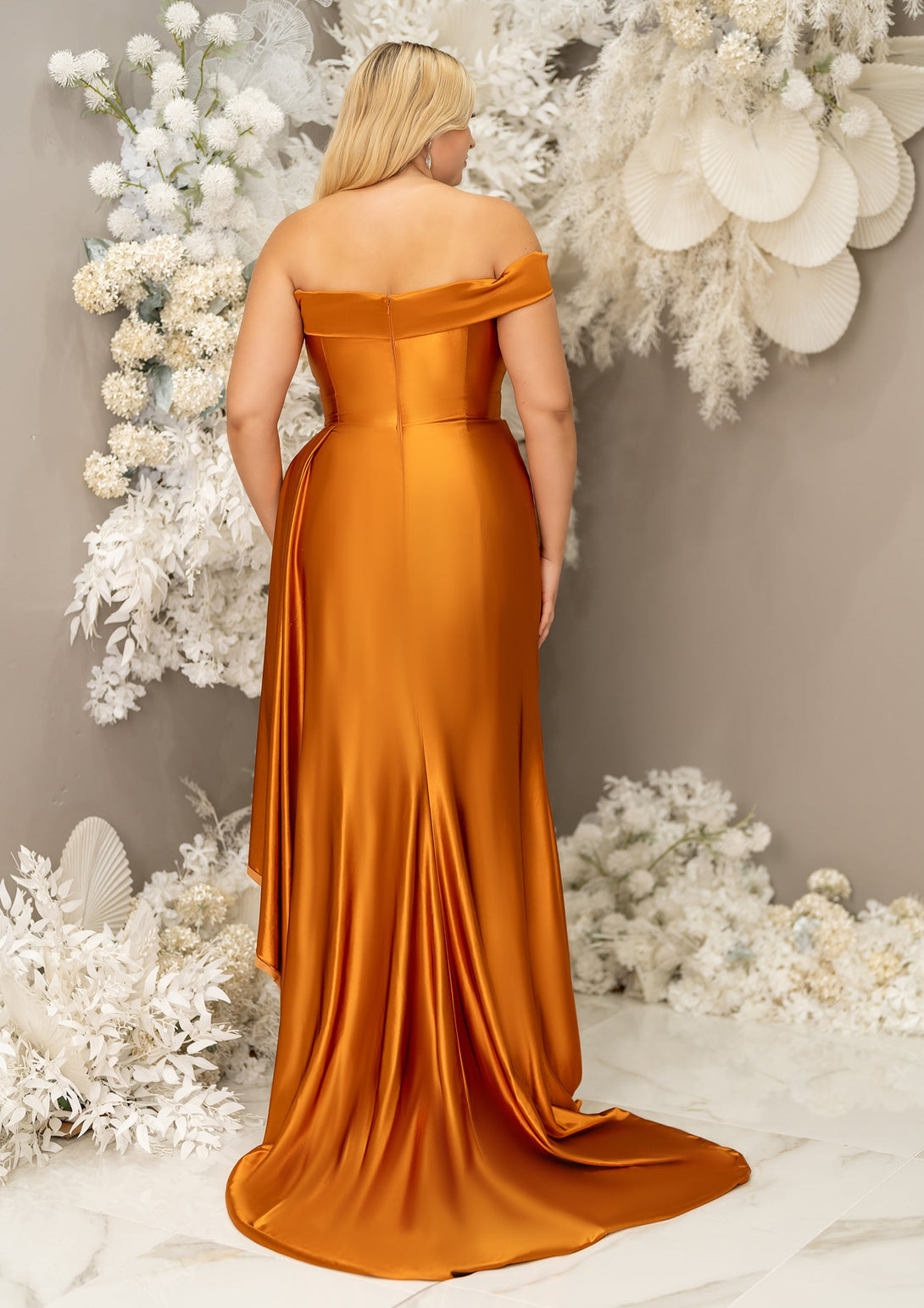 Plus size bridesmaid in asymmetrical fitted stretch satin bridesmaid dress with ruching at the waist and through the skirt and an off-the-shoulder strap. This style is designed to flatter and complement all body types. Shown in burnt orange on a curvy model. Back View