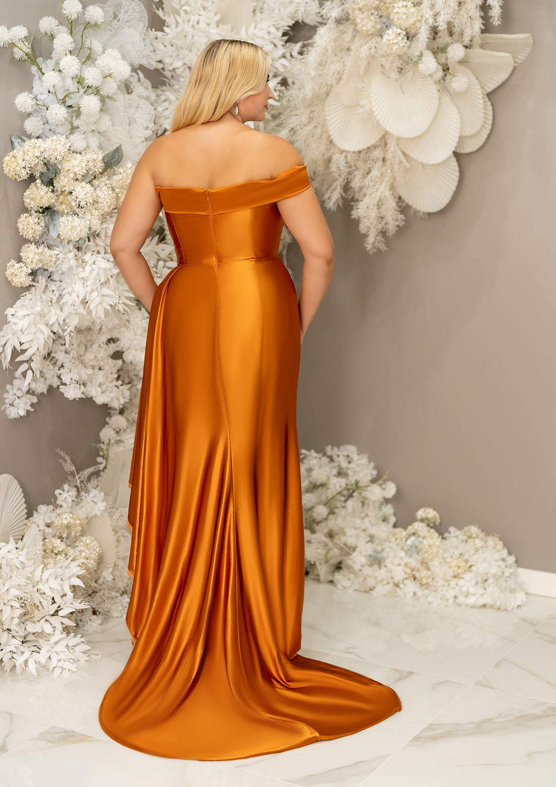 Plus size bridesmaid in asymmetrical fitted stretch satin bridesmaid dress with ruching at the waist and through the skirt and an off-the-shoulder strap. This style is designed to flatter and complement all body types. Shown in burnt orange on a curvy model.